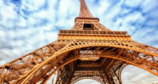 a large metal tower with clouds in the sky with Eiffel Tower in the background