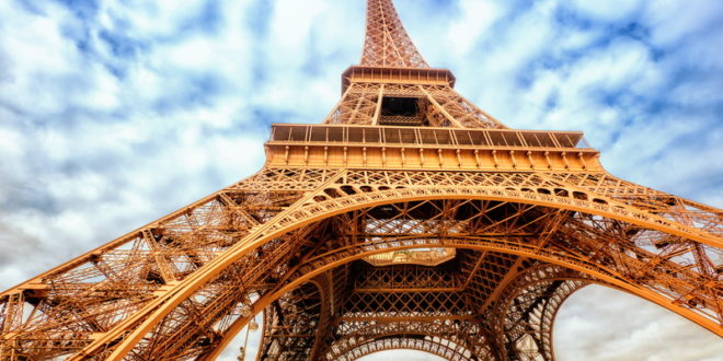a large metal tower with clouds in the sky with Eiffel Tower in the background