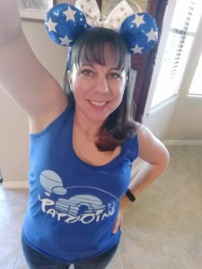 A woman is taking a selfie indoors. She is wearing a blue tank top with a design on it and a headband with blue and white star-patterned Mickey Mouse ears. She is smiling and has one arm raised. The room has light-colored walls and a window with blinds.