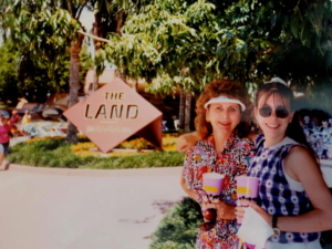The image shows two people standing in front of a sign that reads "The Land." They are both smiling and holding drinks. The person on the left is wearing a floral dress and a visor, while the person on the right is wearing sunglasses and a white shirt. There are trees and greenery in the background.