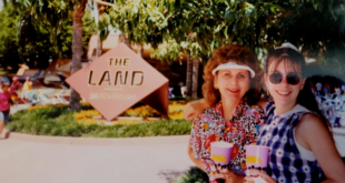 A woman and a young girl are smiling and posing for a photo in front of a sign that reads "The Land." They are holding colorful cups and standing under a tree, with greenery and other people visible in the background. The woman is wearing a colorful patterned shirt and a visor, while the girl is wearing sunglasses and a checkered dress.
