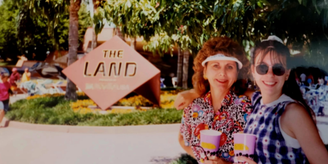 A woman and a young girl are smiling and posing for a photo in front of a sign that reads "The Land." They are holding colorful cups and standing under a tree, with greenery and other people visible in the background. The woman is wearing a colorful patterned shirt and a visor, while the girl is wearing sunglasses and a checkered dress.