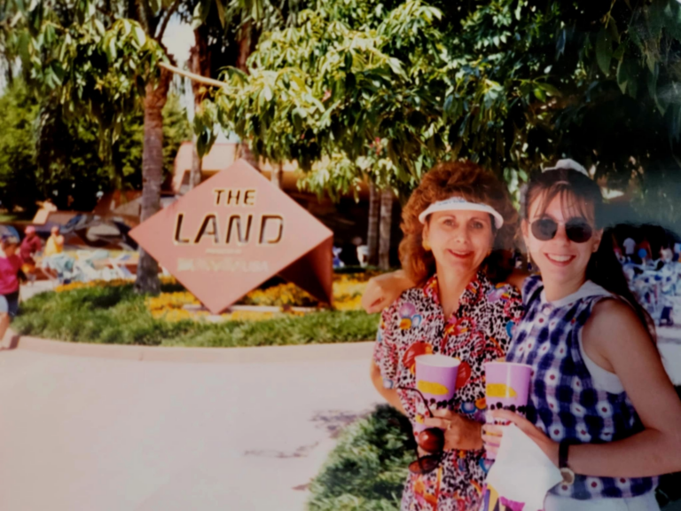 A woman and a young girl are smiling and posing for a photo in front of a sign that reads "The Land." They are holding colorful cups and standing under a tree, with greenery and other people visible in the background. The woman is wearing a colorful patterned shirt and a visor, while the girl is wearing sunglasses and a checkered dress.