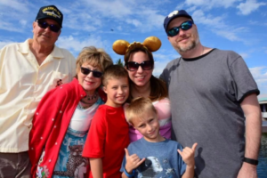 A group of six people, including two children and four adults, are posing together outdoors under a blue sky. One adult is wearing a cap and sunglasses, and another is wearing a headband with large ears. The group is smiling, and one child is making a hand gesture. They appear to be enjoying a sunny day.