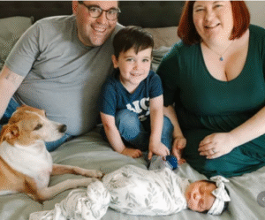 A family is sitting on a bed, smiling at the camera. There is a man and a woman, a young boy, and a newborn baby wrapped in a patterned blanket. A dog is also lying on the bed next to the family. The atmosphere is warm and joyful.