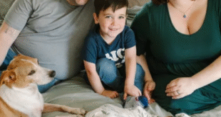 A family is sitting on a bed, smiling at the camera. There is a man and a woman, a young boy, and a newborn baby wrapped in a patterned blanket. A dog is also lying on the bed next to the family. The atmosphere is warm and joyful.