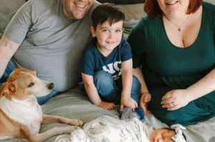 A family is sitting on a bed, smiling at the camera. There is a man and a woman, a young boy, and a newborn baby wrapped in a patterned blanket. A dog is also lying on the bed next to the family. The atmosphere is warm and joyful.